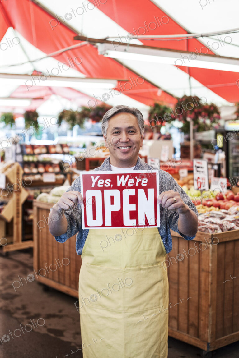 Smiling man wearing apron at a fruit and vegetable market, holding aloft Open sign..