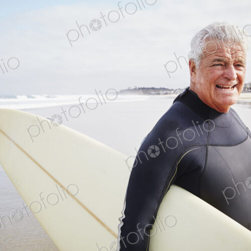 Smiling senior man standing on a beach, wearing a wetsuit and carrying a surfboard. Smiling senior man standing on a beach, wearing a wetsuit and carrying a surfboard.