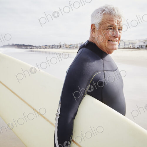 Smiling senior man standing on a beach, wearing a wetsuit and carrying a surfboard. Smiling senior man standing on a beach, wearing a wetsuit and carrying a surfboard.