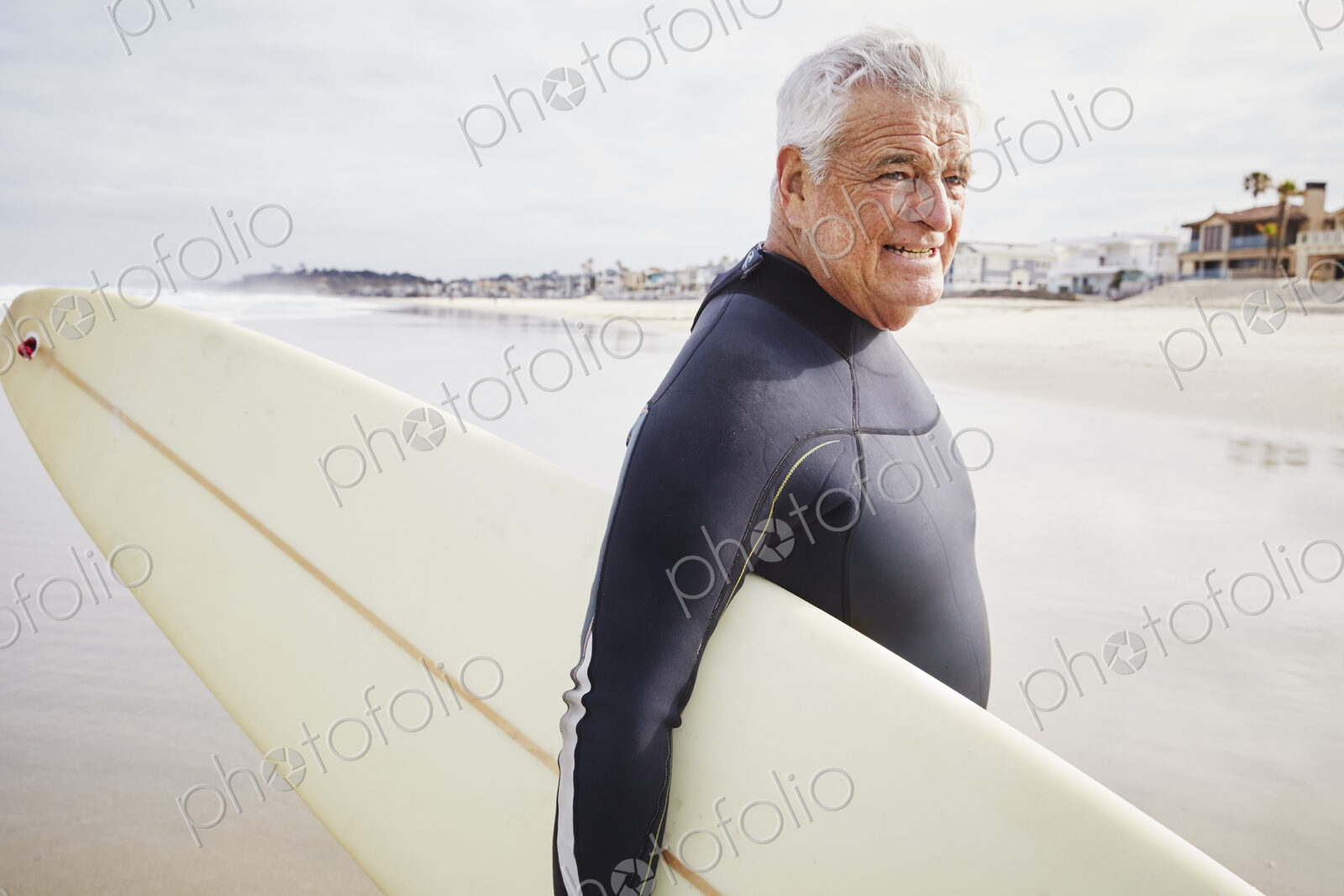 Smiling senior man standing on a beach, wearing a wetsuit and carrying a surfboard.