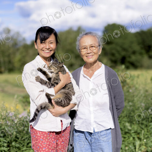 Smiling woman holding grey cat standing outdoors, with an elderly woman, smiling at… Smiling woman holding grey cat standing outdoors, with an elderly woman, smiling at…