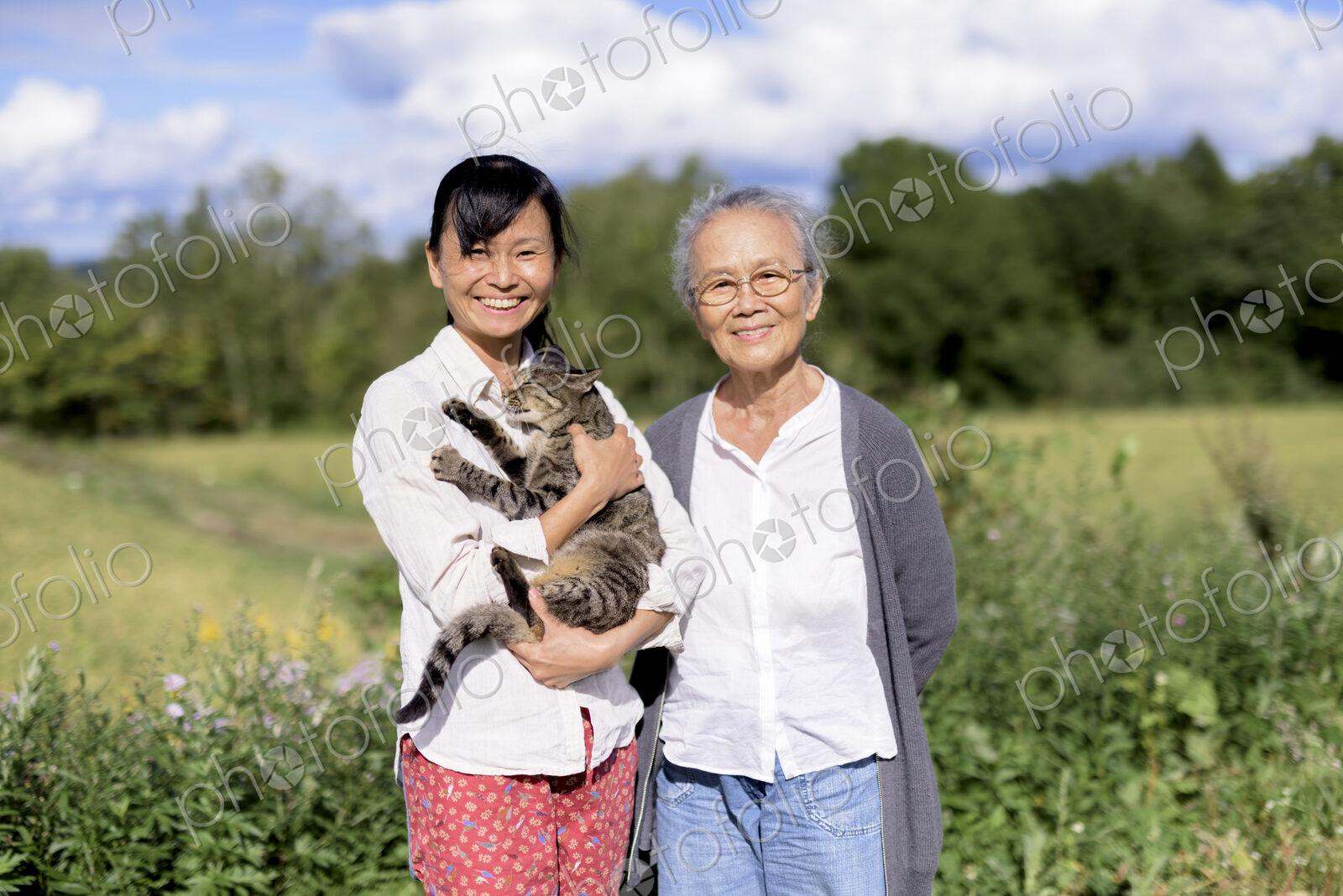 Smiling woman holding grey cat standing outdoors, with an elderly woman, smiling at…