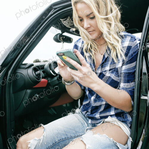Smiling woman in ripped jeans sitting in car, holding mobile phone, texting.