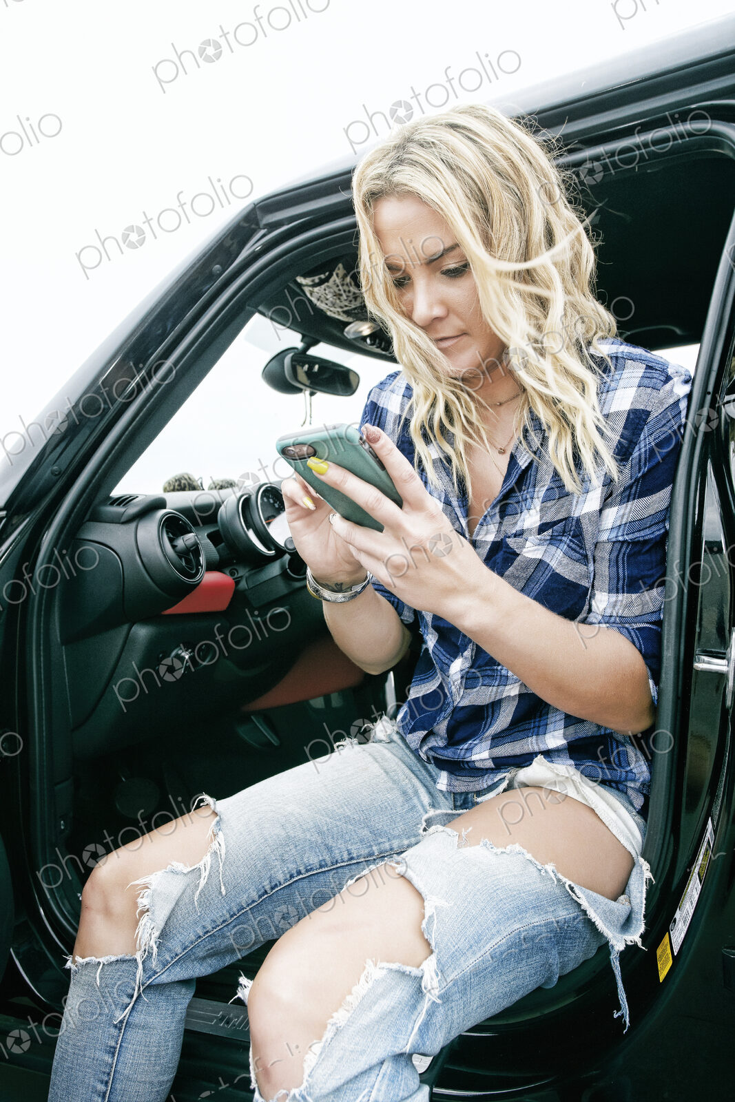 Smiling woman in ripped jeans sitting in car, holding mobile phone, texting.
