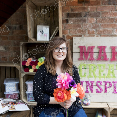 Smiling woman sitting in a workshop, holding bunch of colourful handcrafted fabric…