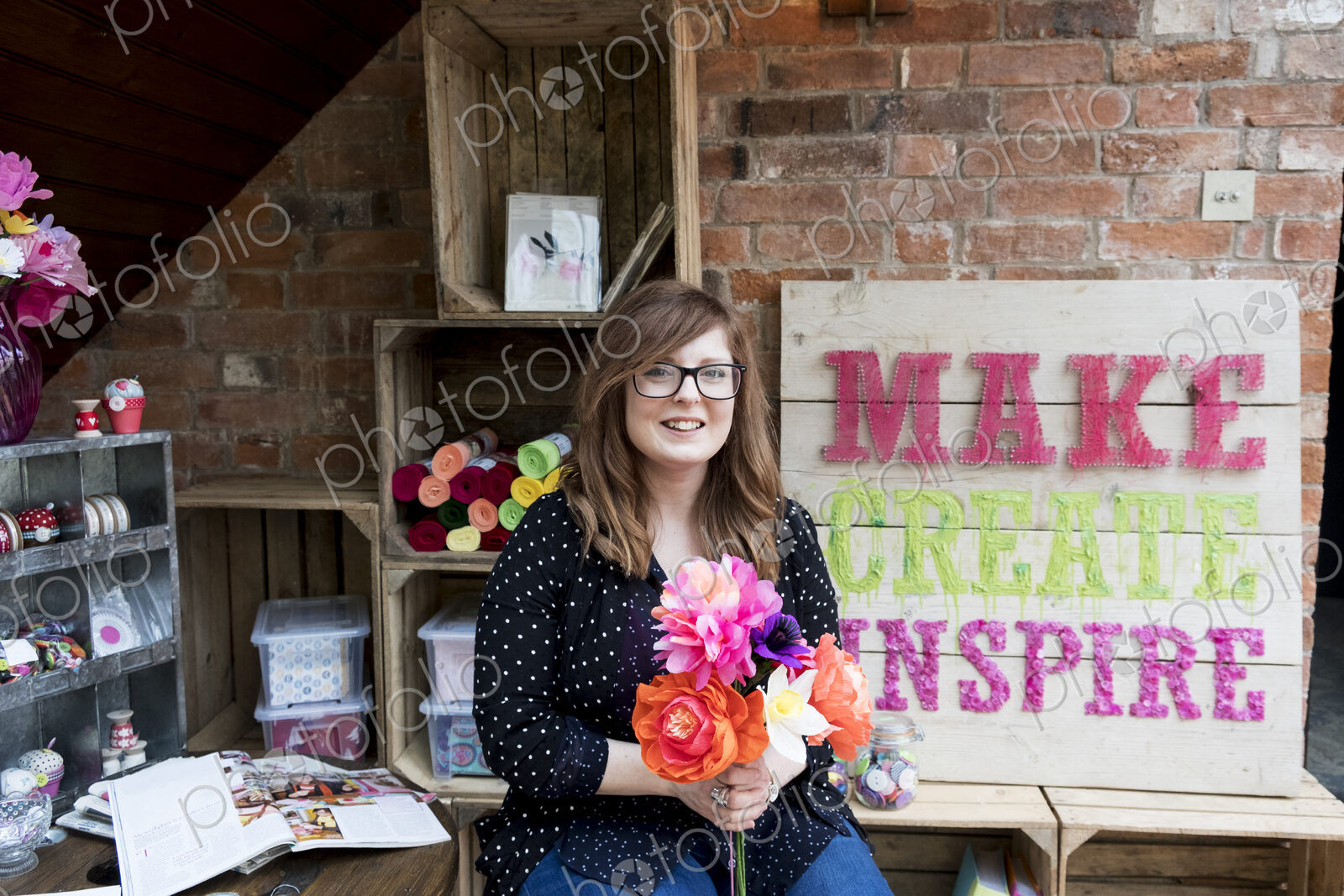 Smiling woman sitting in a workshop, holding bunch of colourful handcrafted fabric…