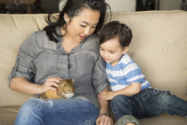 Smiling woman sitting on a sofa, a guinea pig sitting on her lap, her young son watching.