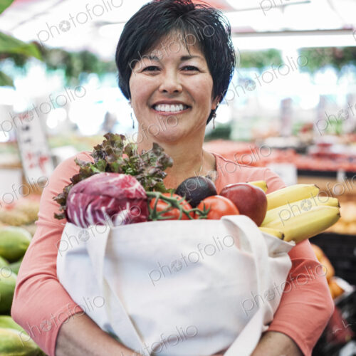 Smiling woman standing in a food and vegetable market, holding shopping bag with fresh…
