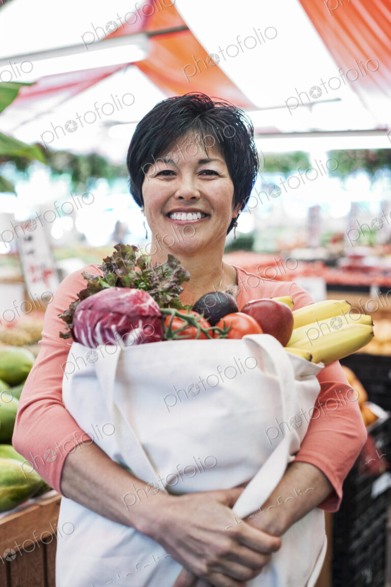 Smiling woman standing in a food and vegetable market, holding shopping bag with fresh…