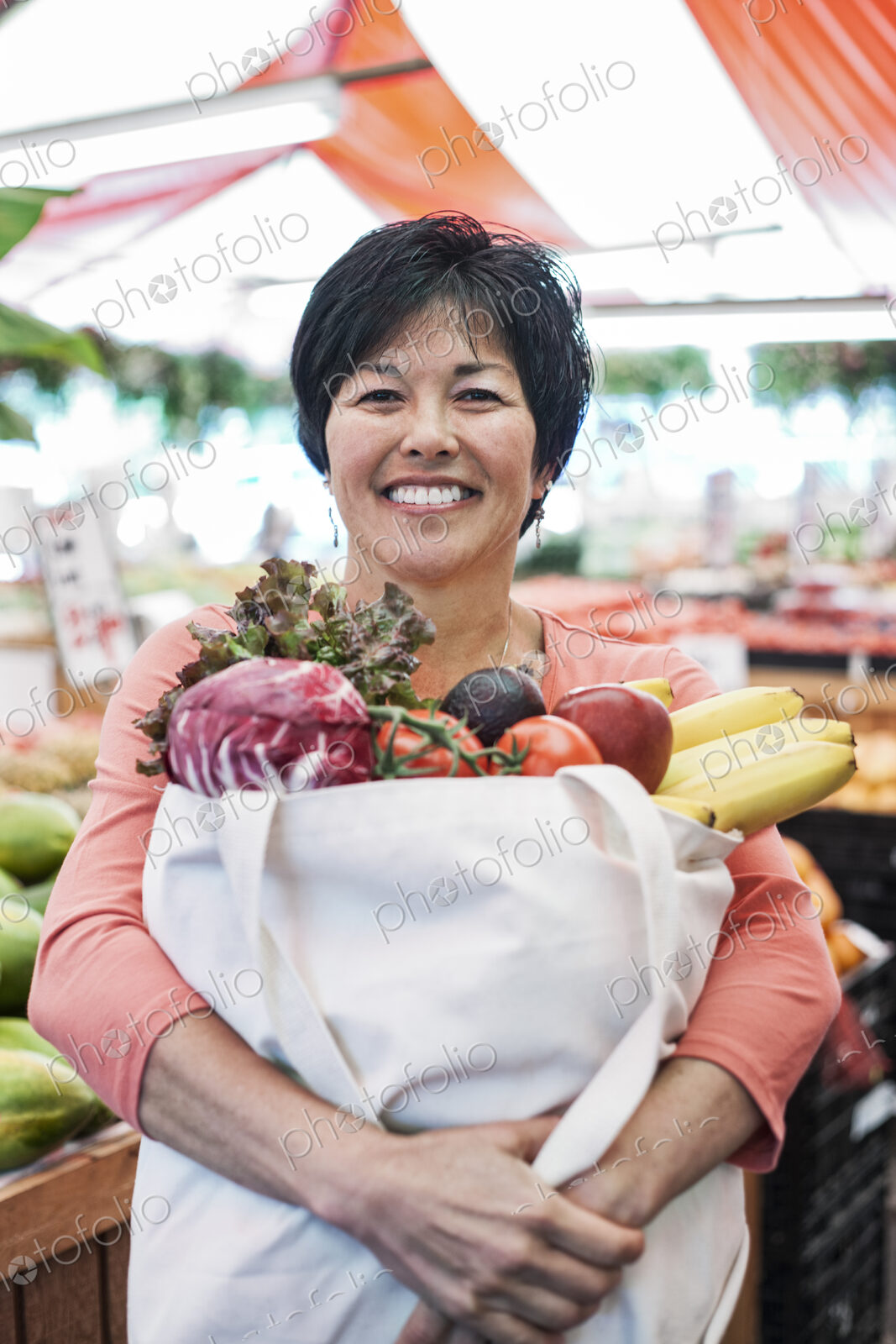 Smiling woman standing in a food and vegetable market, holding shopping bag with fresh…