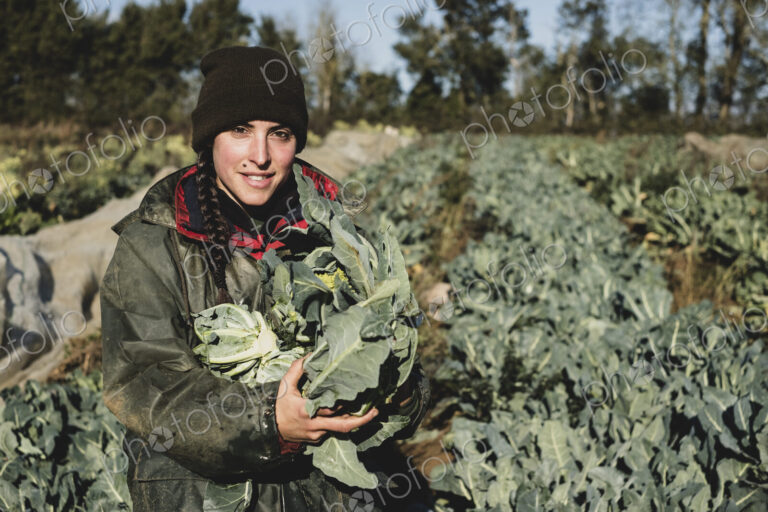 Smiling woman standing in field, holding blue crate with freshly harvested…