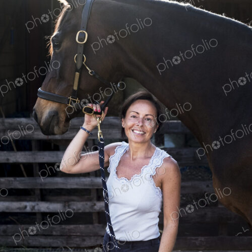Smiling woman standing outdoor at stable, holding brown horse by rein. Smiling woman standing outdoor at stable, holding brown horse by rein.