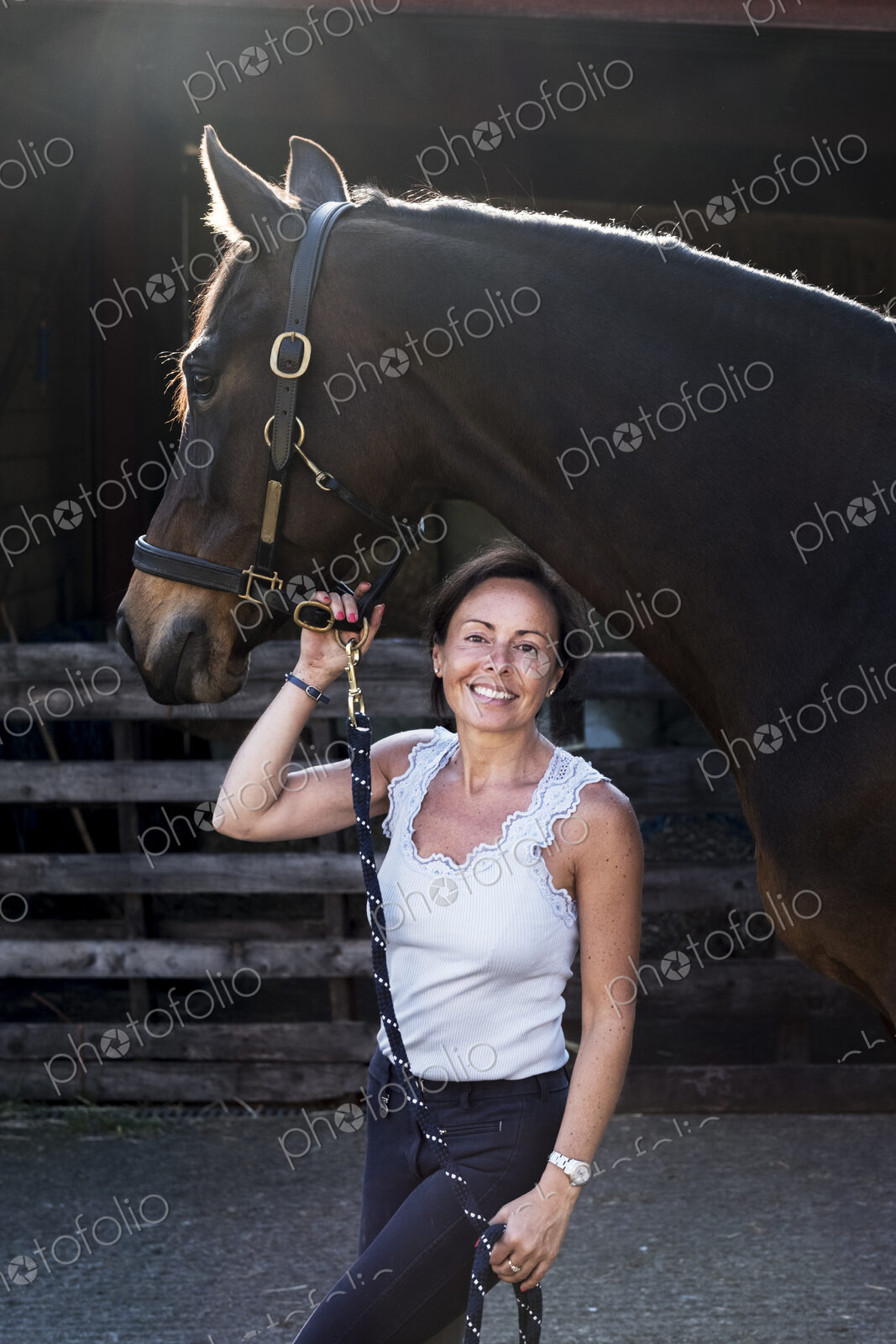 Smiling woman standing outdoor at stable, holding brown horse by rein.
