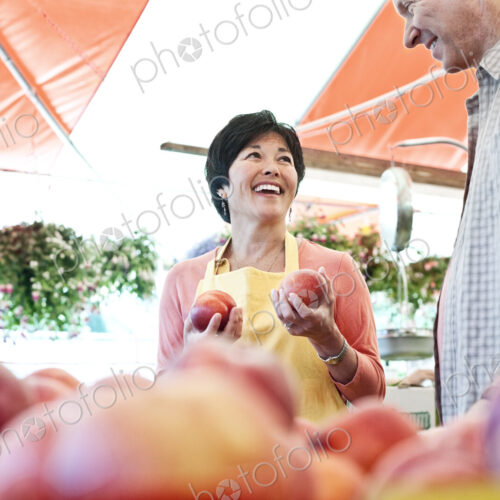 Smiling woman wearing apron and man standing at stall with fresh peaches at a fruit… Smiling woman wearing apron and man standing at stall with fresh peaches at a fruit…
