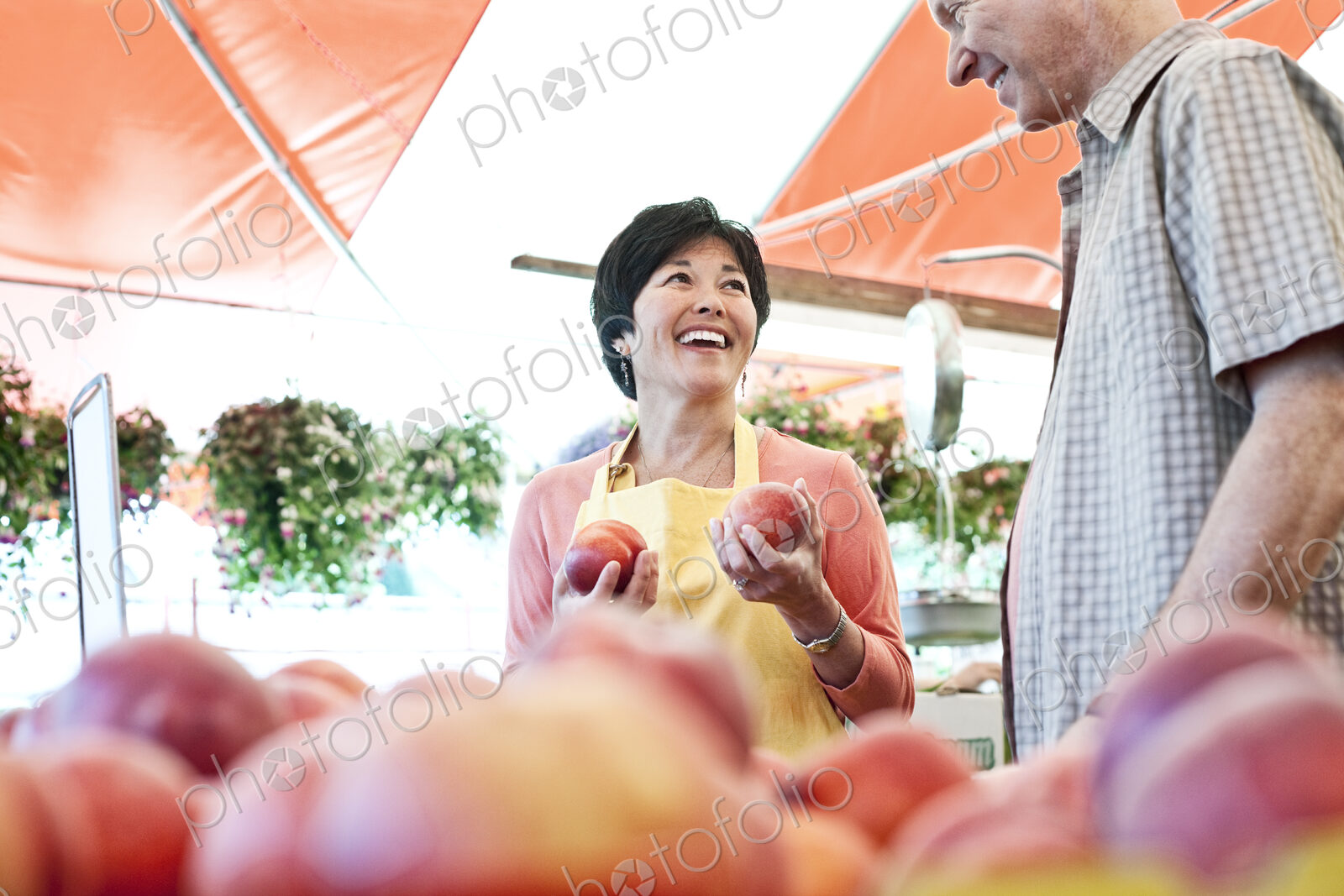 Smiling woman wearing apron and man standing at stall with fresh peaches at a fruit…
