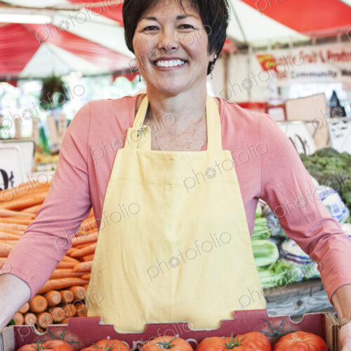 Smiling woman wearing apron holding large tray of fresh tomatoes at a fruit and… Smiling woman wearing apron holding large tray of fresh tomatoes at a fruit and…