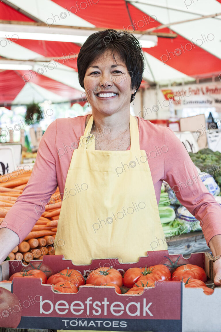 Smiling woman wearing apron holding large tray of fresh tomatoes at a fruit and…