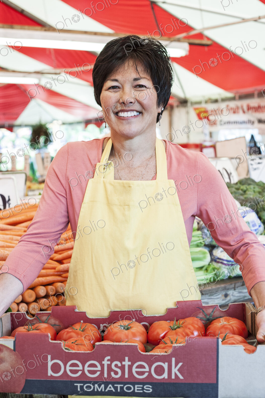 Smiling woman wearing apron holding large tray of fresh tomatoes at a fruit and…