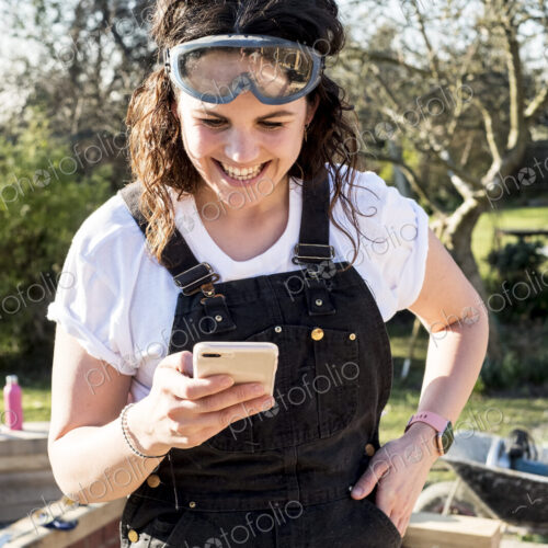 Smiling woman wearing dungarees and protective goggles checking her mobile phone. Smiling woman wearing dungarees and protective goggles checking her mobile phone.