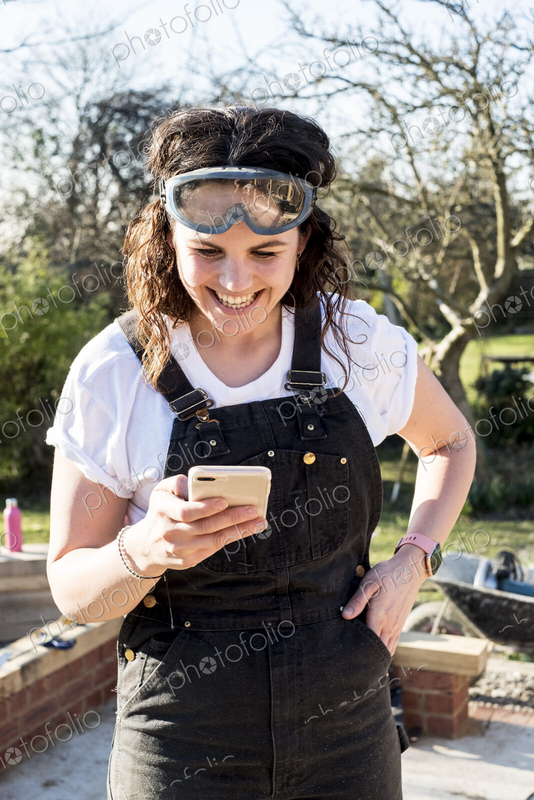 Smiling woman wearing dungarees and protective goggles checking her mobile phone.