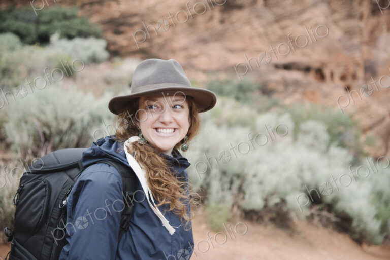 Smiling woman with auburn hair, wearing a hat and carrying a backpack, hiking in a canyon.