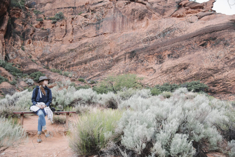 Smiling woman with auburn hair, wearing a hat sitting on a bench in a canyon.