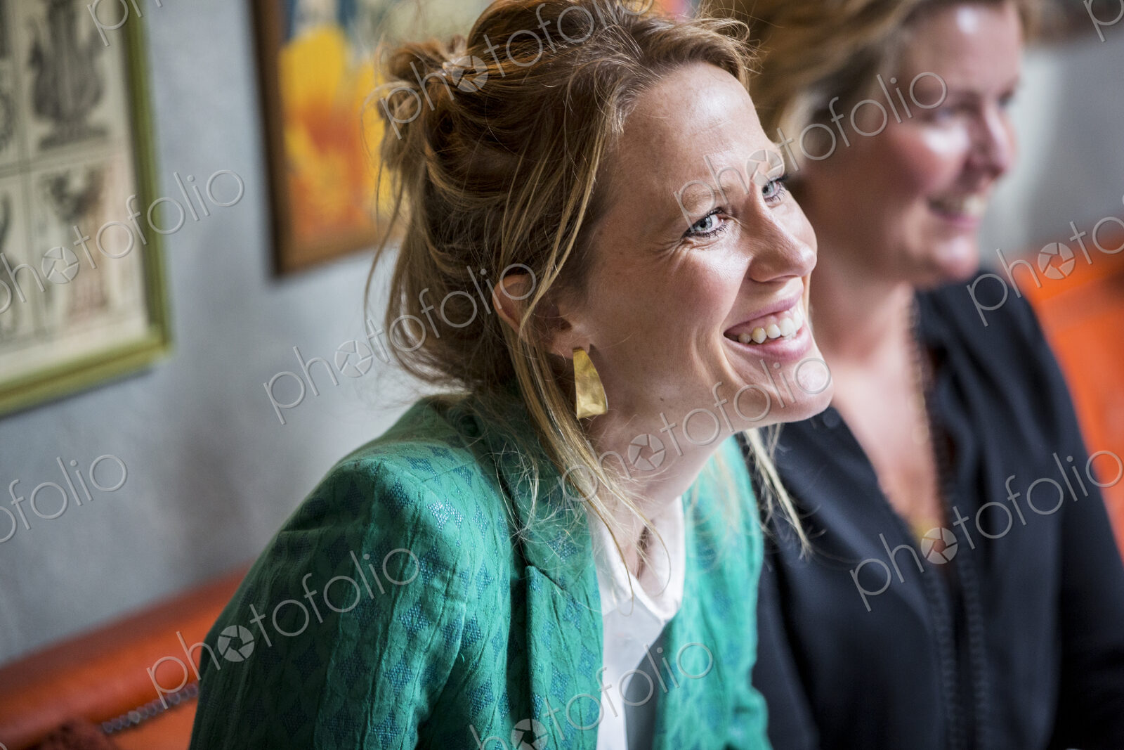 Smiling woman with blond hair wearing golden earrings and green jacket sitting at table.