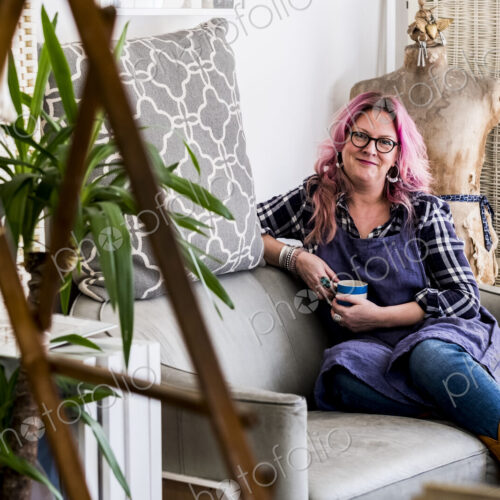 Smiling woman with long blond wavy hair with pink streaks sitting on a sofa, looking…