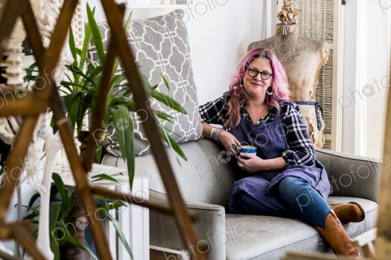 Smiling woman with long blond wavy hair with pink streaks sitting on a sofa, looking…