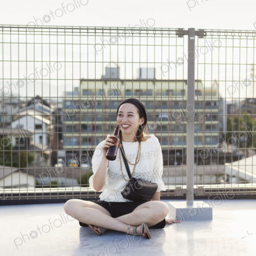 Smiling young Japanese woman sitting on a rooftop in an urban setting, drinking beer. Smiling young Japanese woman sitting on a rooftop in an urban setting, drinking beer.