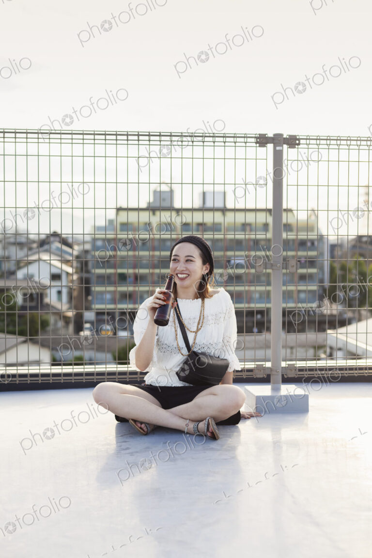 Smiling young Japanese woman sitting on a rooftop in an urban setting, drinking beer.