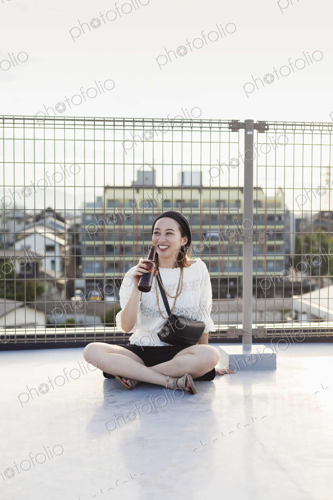 Smiling young Japanese woman sitting on a rooftop in an urban setting, drinking beer.