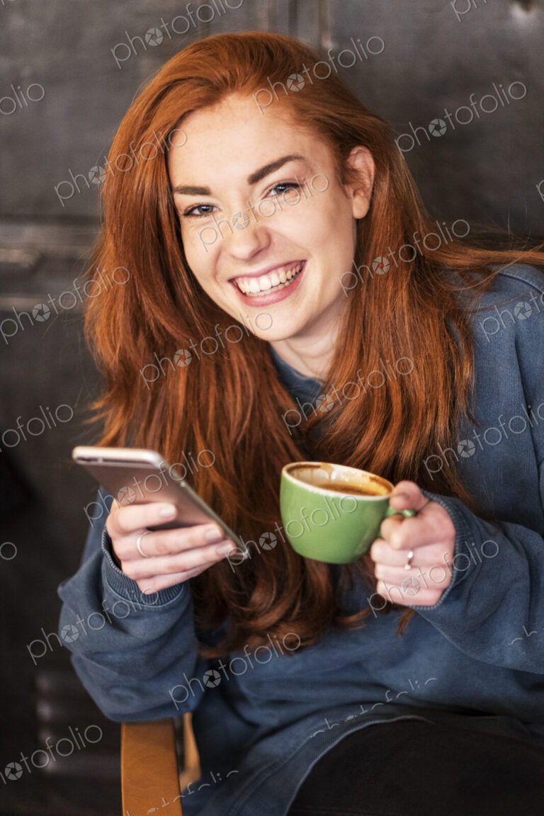Smiling young woman holding mobile phone and coffee, working from home