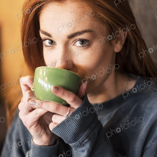 Smiling young woman with long red hair holding cup of coffee, looking at camera.