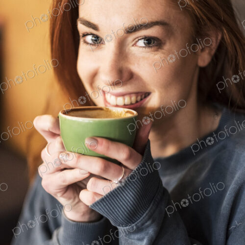 Smiling young woman with long red hair holding cup of coffee, looking at camera.