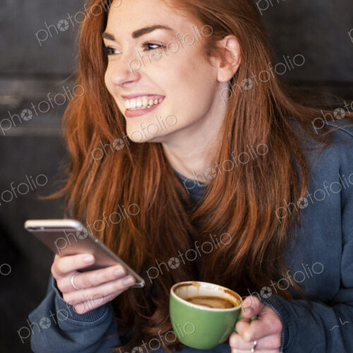 Smiling young woman with long red hair sitting at table, holding mobile phone and cup…