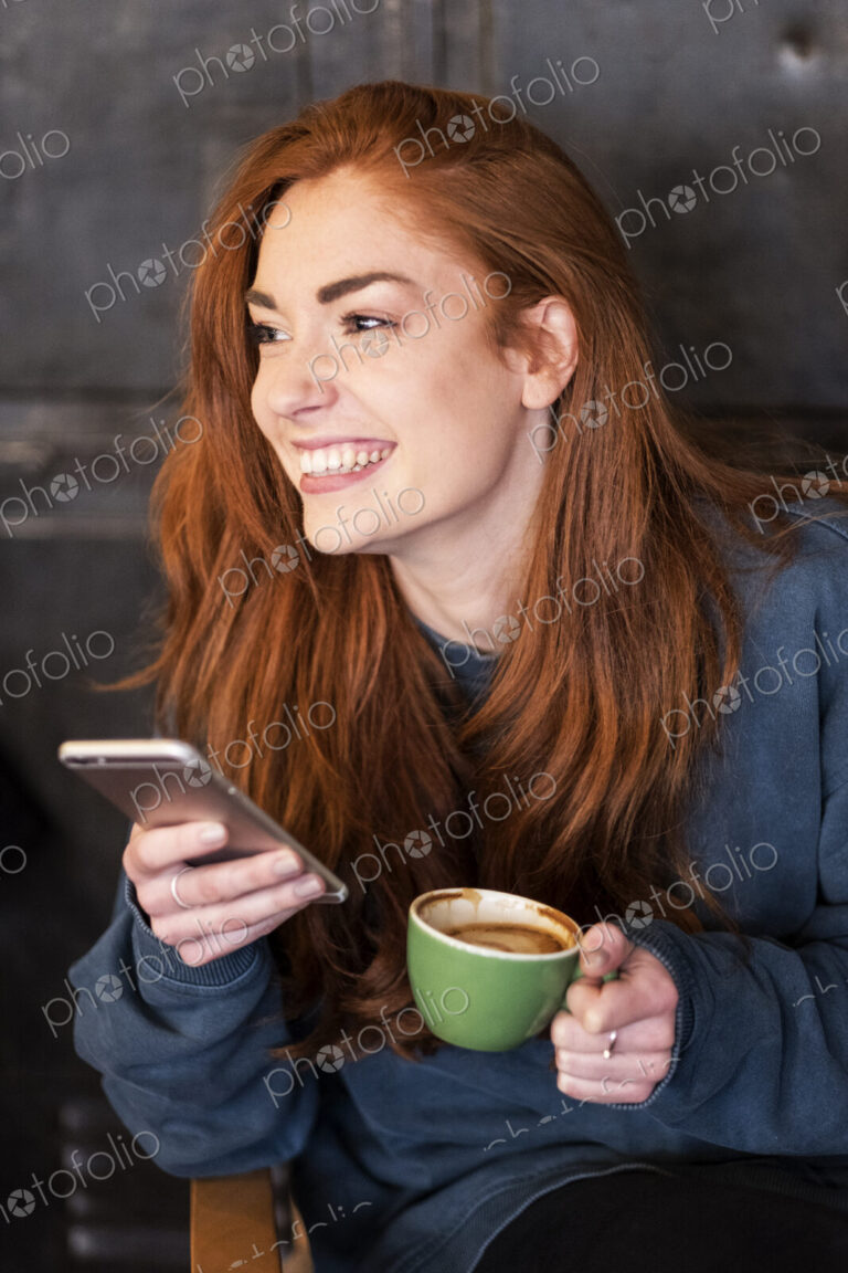 Smiling young woman with long red hair sitting at table, holding mobile phone and cup…