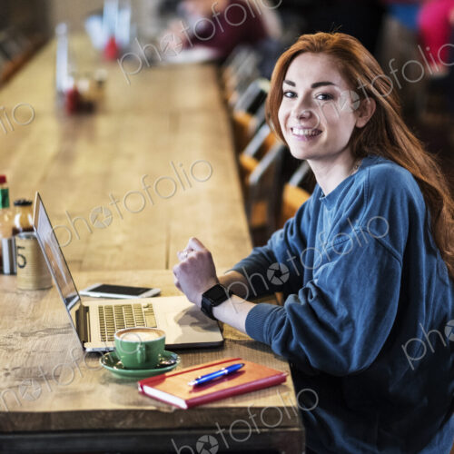 Smiling young woman with long red hair sitting at table, working on laptop computer. Smiling young woman with long red hair sitting at table, working on laptop computer.