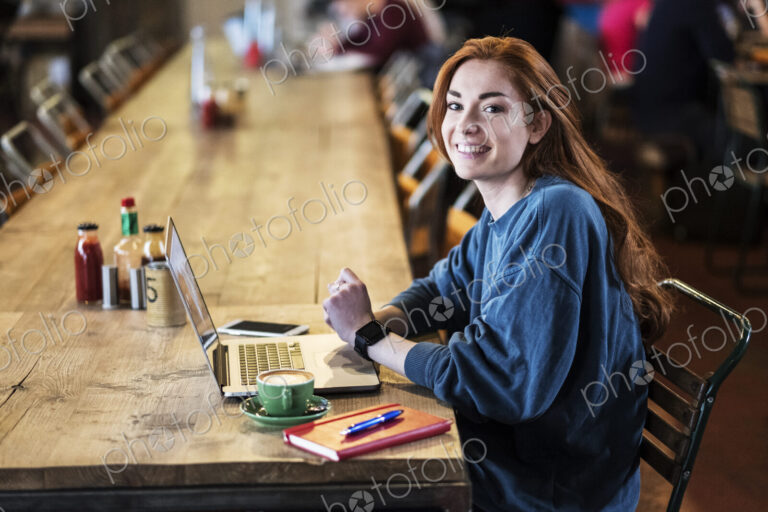 Smiling young woman with long red hair sitting at table, working on laptop computer.