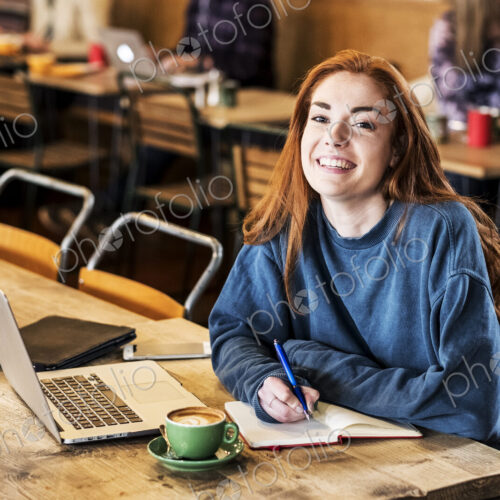Smiling young woman with long red hair sitting at table, working on laptop computer. Smiling young woman with long red hair sitting at table, working on laptop computer.