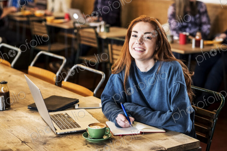 Smiling young woman with long red hair sitting at table, working on laptop computer.