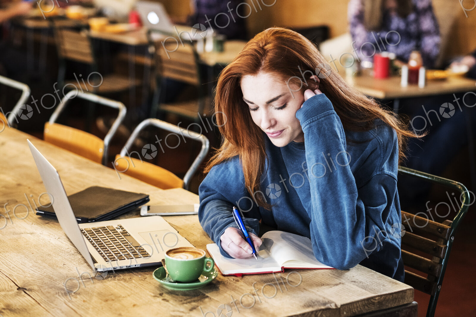 Smiling young woman with long red hair sitting at table, working on laptop computer.