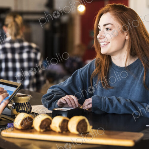 Smiling young woman with long red hair standing at a counter in a restaurant, paying…