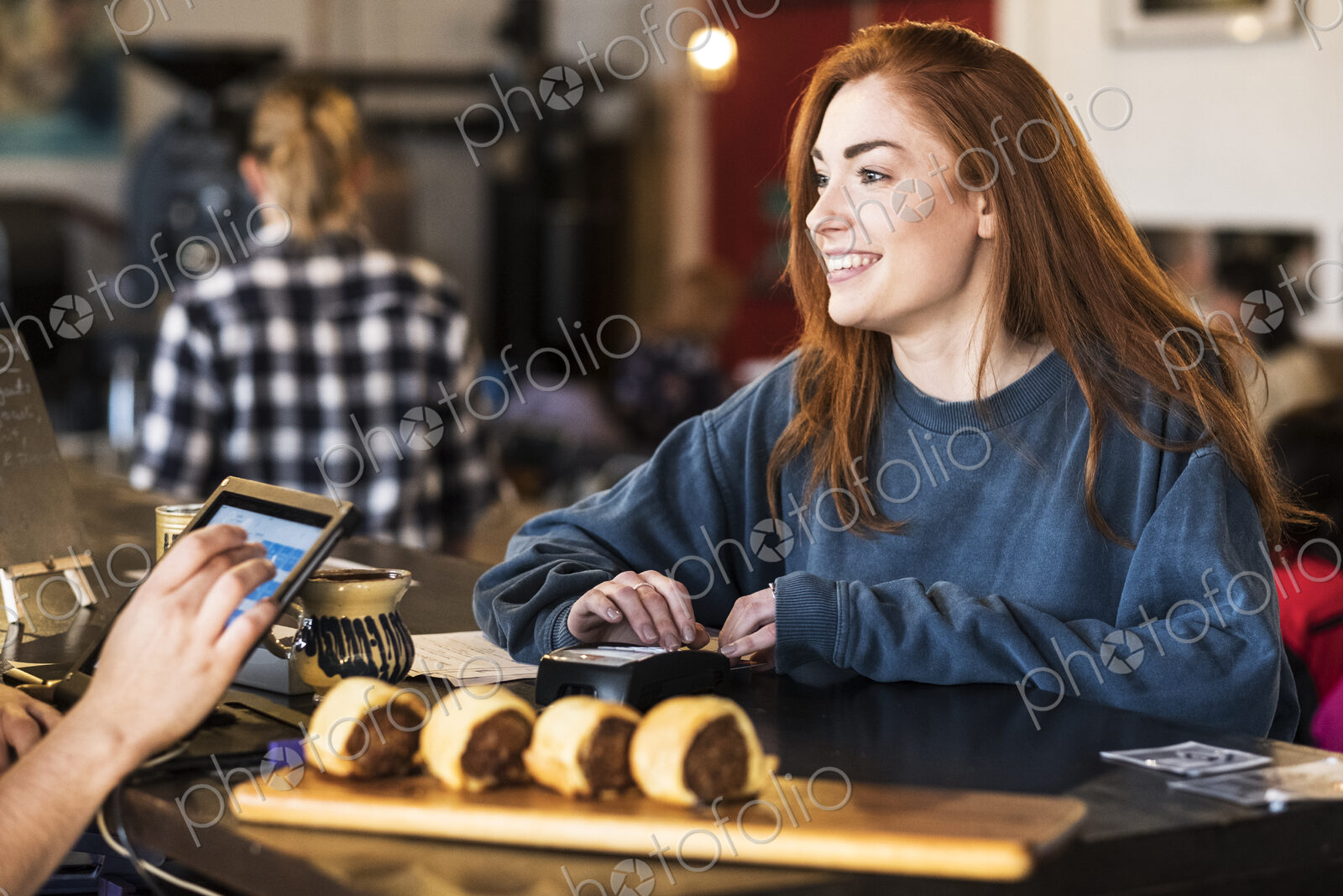 Smiling young woman with long red hair standing at a counter in a restaurant, paying…