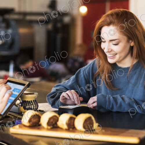 Smiling young woman with long red hair standing at a counter in a restaurant, paying…