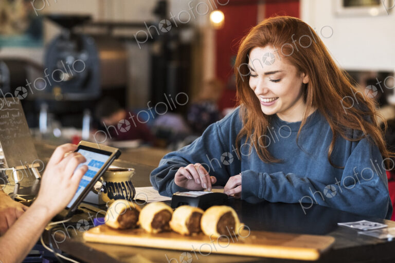 Smiling young woman with long red hair standing at a counter in a restaurant, paying…