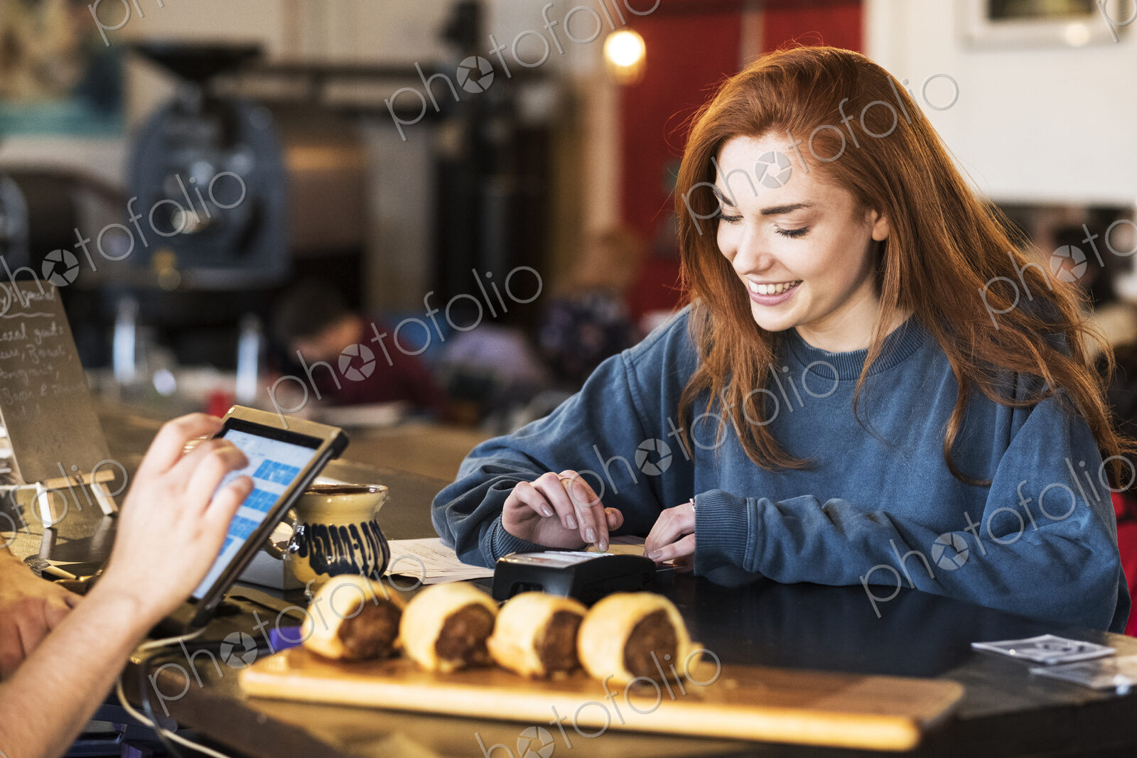 Smiling young woman with long red hair standing at a counter in a restaurant, paying…