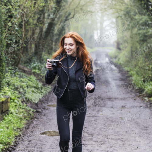 Smiling young woman with long red hair walking along forest path, taking pictures with…