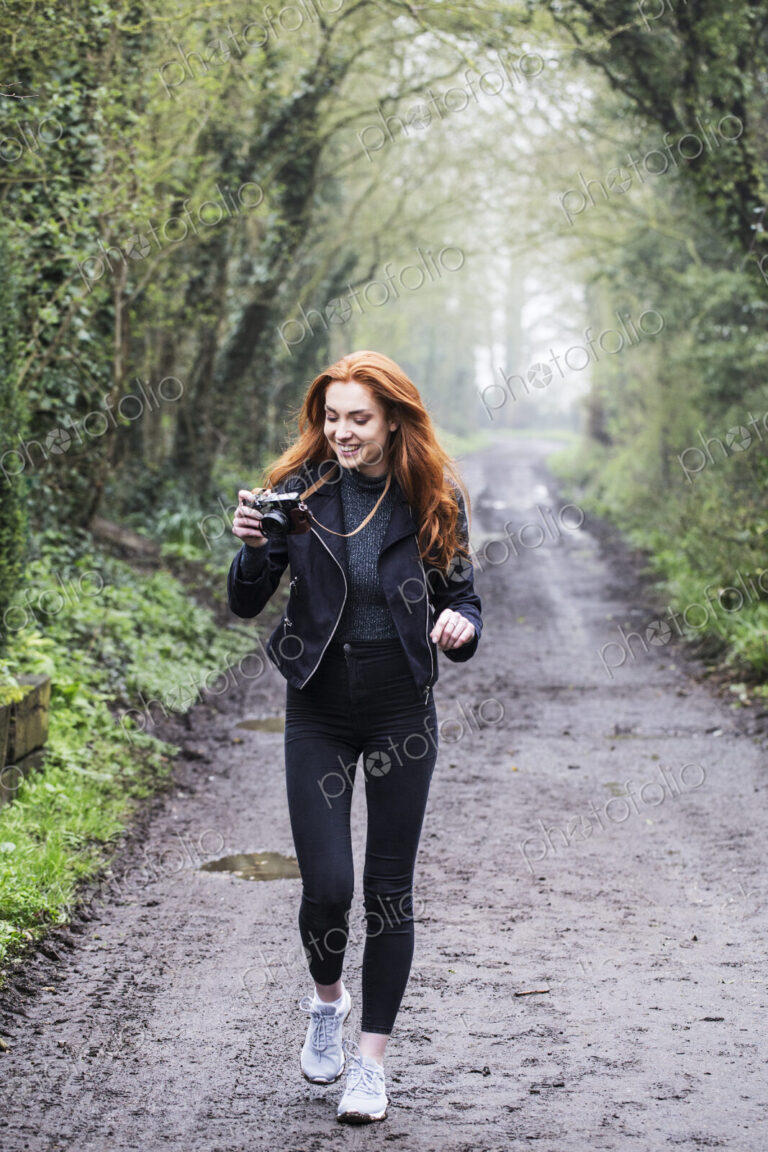 Smiling young woman with long red hair walking along forest path, taking pictures with…