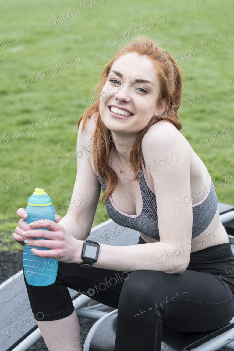 Smiling young woman with long red hair wearing sportswear, exercising outdoors,…